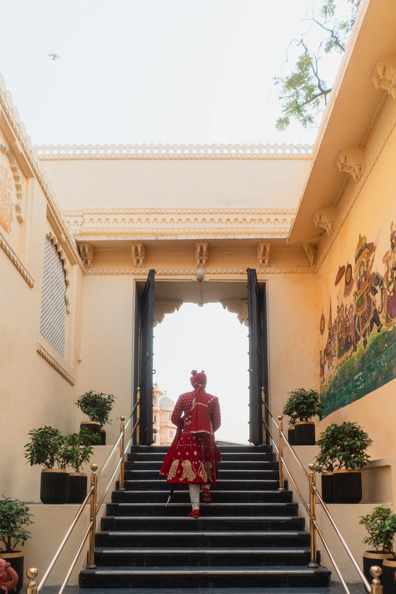 A person dressed in traditional Indian attire climbing a staircase at a historic building with intricate architecture, potted plants, and a colorful mural.