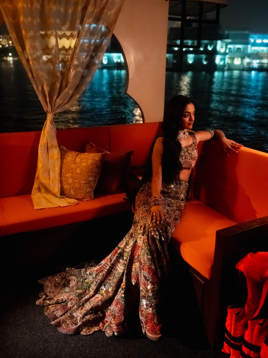 A woman dressed in a heavily embellished traditional Indian outfit with a long, patterned flowing skirt and matching top, sitting on an orange cushioned seat near water at night with city lights in the background.