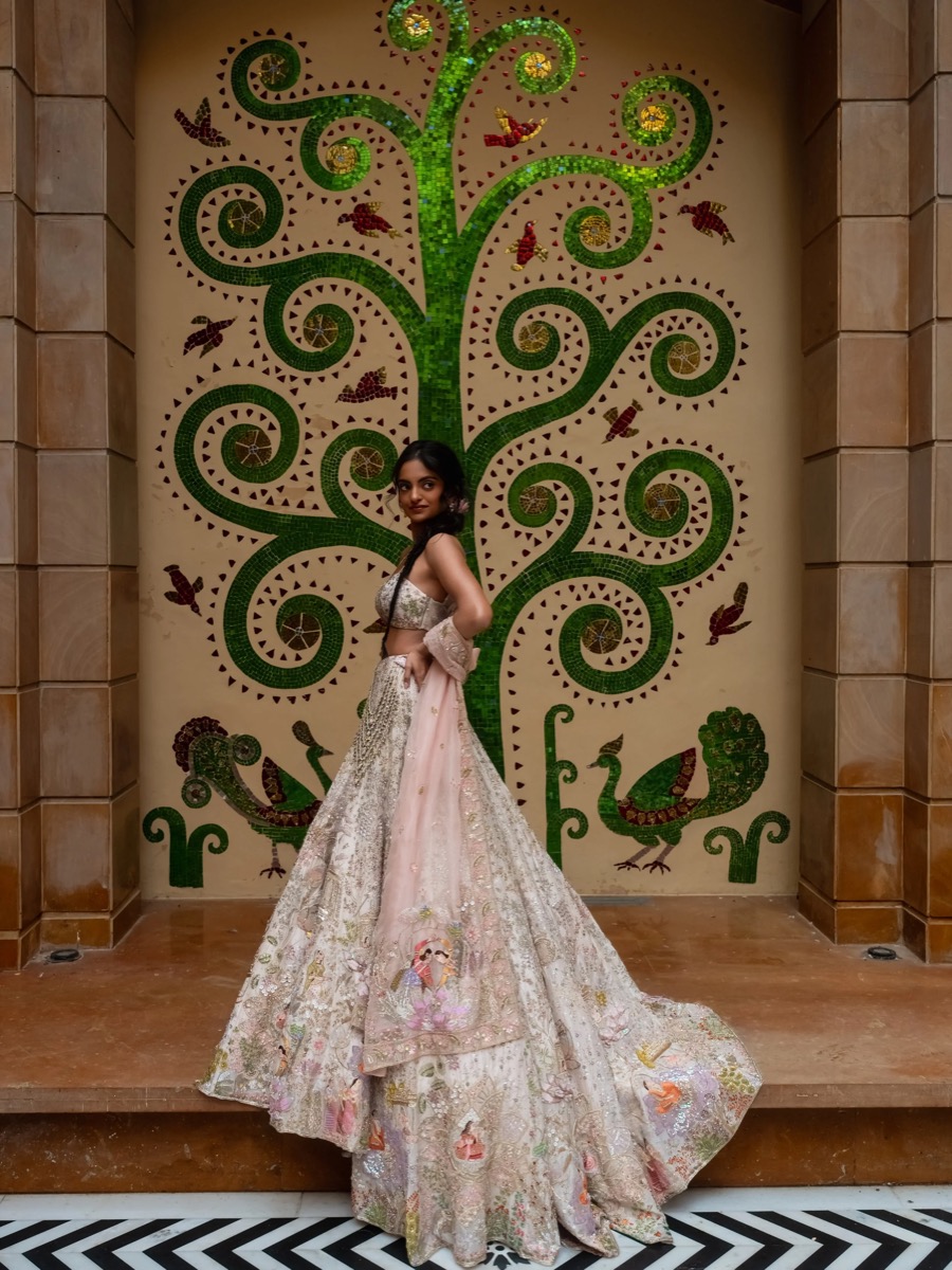 A woman in traditional Indian attire standing in front of a decorative mosaic wall with a green swirling tree, birds, and peacocks.