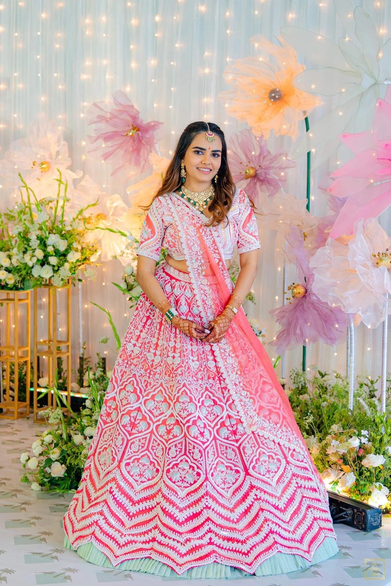 A woman in traditional Indian attire standing in front of a floral backdrop with lights. She is wearing a pink and white embroidered lehenga, jewelry including necklaces, earrings, and a maang tikka, and has Mehndi on her hands.