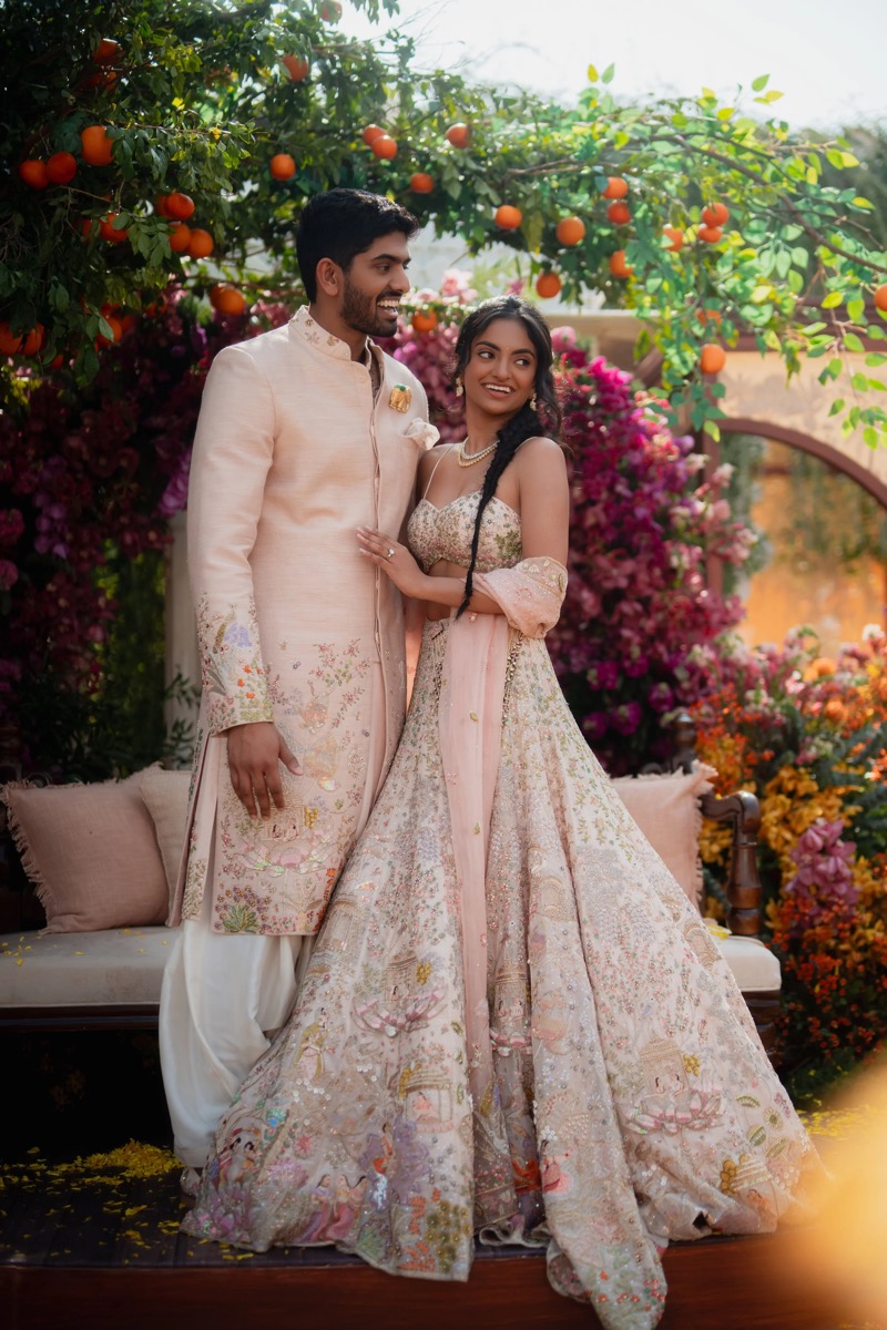 A couple in traditional Indian wedding attire standing outdoors, smiling and embracing among colorful flowers and orange trees.