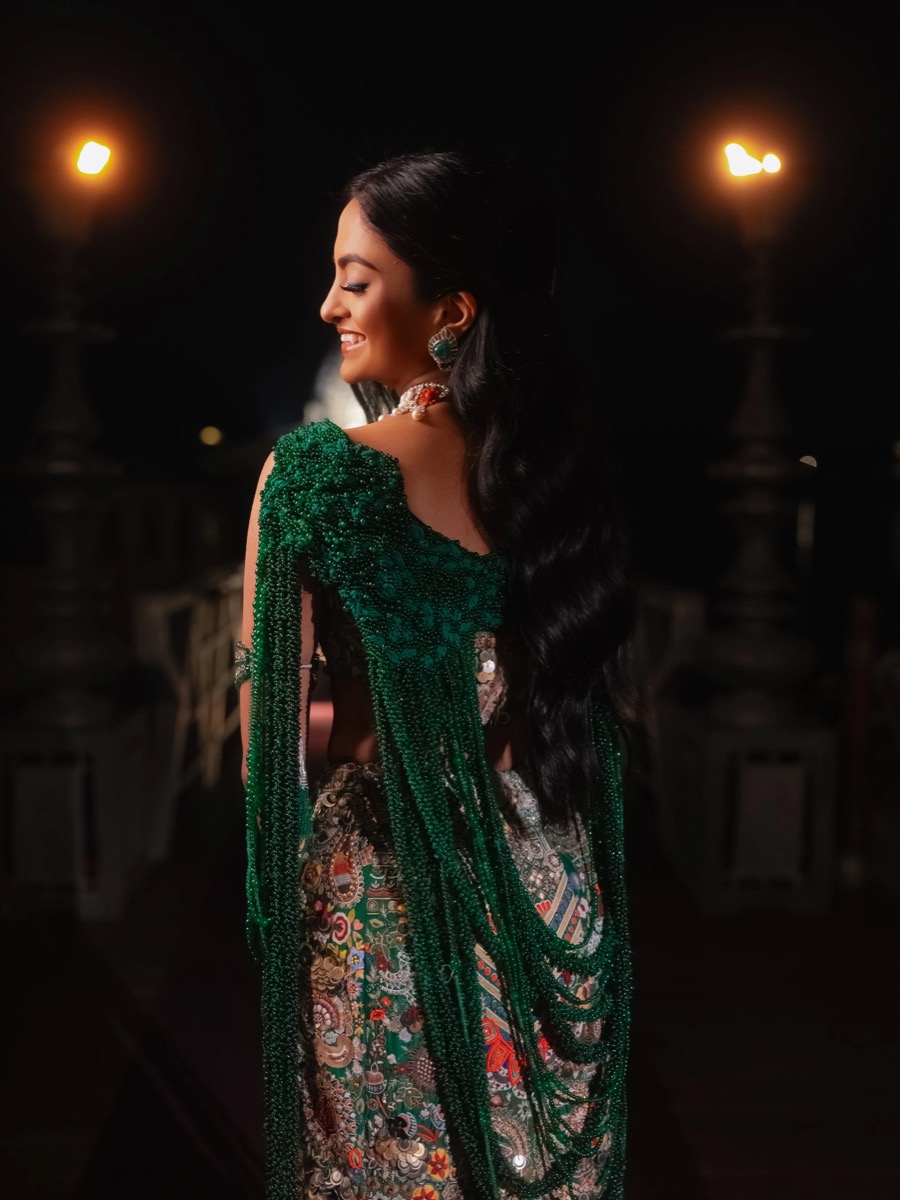 Side view of a smiling woman with long black hair styled in loose waves, wearing a green beaded top and a patterned skirt, decorated with jewelry, in a nighttime outdoor setting illuminated by streetlights.