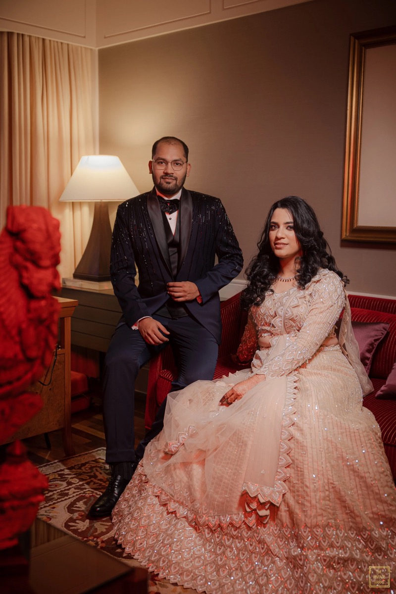 A man in a black tuxedo with a bowtie and a woman in a cream-colored, heavily embroidered dress sitting on a red sofa in a warmly lit room.