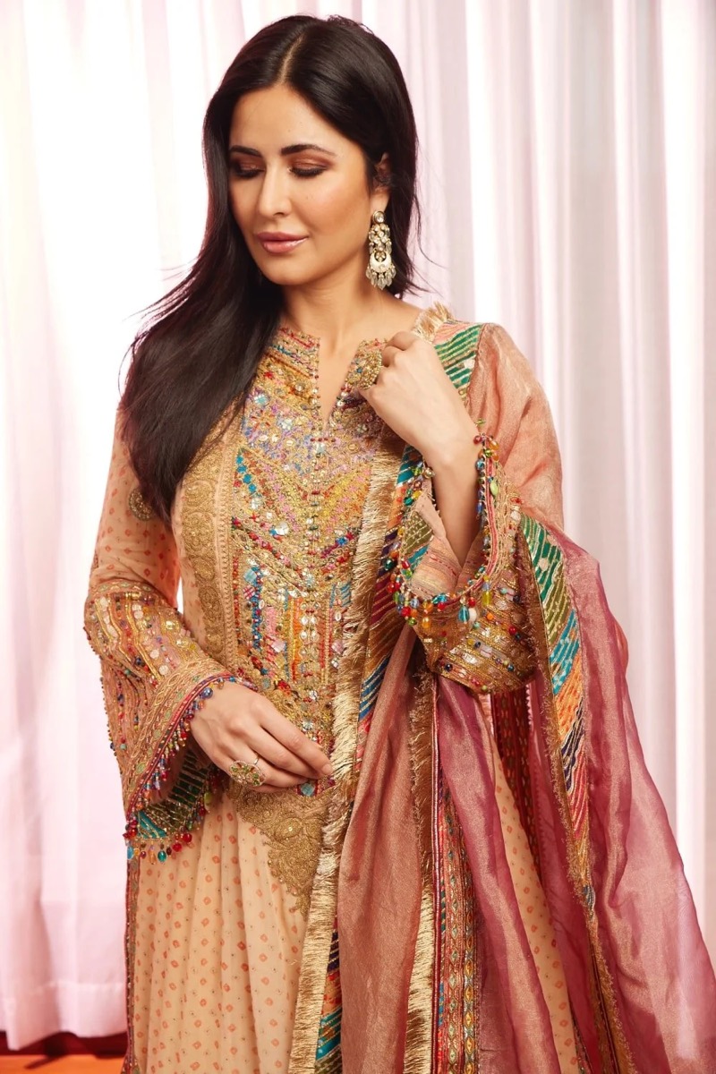 A woman with long dark hair, wearing a traditional embroidered dress with multicolored beads and gold accents, posing indoors with a soft pink curtain background.