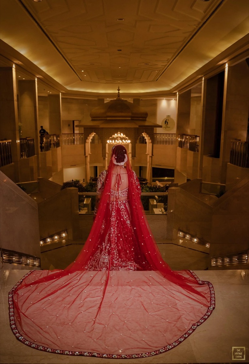 A bride dressed in a red and gold traditional wedding gown with a long veil, standing at the top of a staircase in an elegant indoor setting, possibly a hotel lobby or wedding venue.